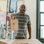 A smiling entrepreneur holding a paper bag with stickers in an artistic setting, showcasing small business creativity.