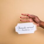 Close-up of a woman's hand holding a 'Female Owned Business' sign on a beige background.